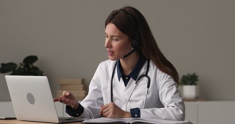 Focused young female doctor general practitioner wearing wireless headset with microphone, giving online video call consultation on computer or taking part in medical educational virtual event.
