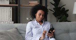 Smiling attractive millennial african american woman using cellphone, playing mobile games, communicating in social network, shopping in internet store, entertaining online resting on cozy couch.