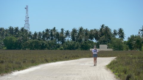 A girl in a medical mask walks along a sandy road towards us, against the backdrop of palm trees and a telecamunactivation tower. A girl in denim blue shorts comes close. Young woman on vacation. 