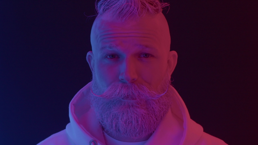 Portrait of adult bearded serious male looking at camera in modern colorful neon lighting at black background indoor close up. Amazed face expression, surprised human emotion of cool posing senior man Royalty-Free Stock Footage #1077212273