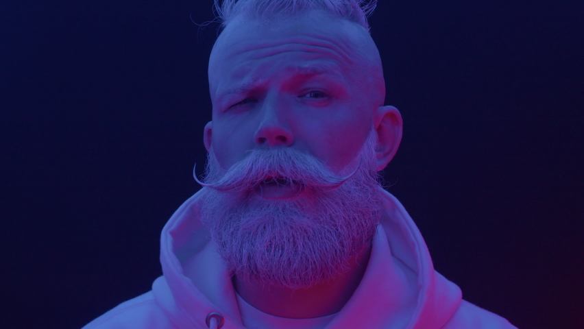 Portrait of adult bearded serious male looking at camera in modern colorful neon lighting at black background indoor close up. Amazed face expression, surprised human emotion of cool posing senior man | Shutterstock HD Video #1077212273