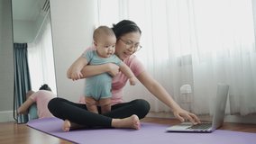 Healthy Asian Young mother embracing or hugging her baby boy while having online yoga class on laptop at home. Sporty Mom playing with her son while exercising on yoga mat at the floor together.