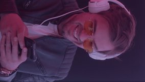 Portrait of stylish man dancing to listening trendy music wearing headphones using smart phone app, enjoying funny relax in colorful dynamic neon illumination indoors close up. Vertical video format