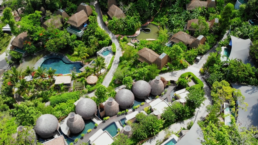 Aerial top down view of beautiful resort with swimming pool and hut shape themed houses and villa surrounded by trees and greenery