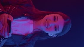 Portrait of young happy smiling asian girl listening modern music wearing earphones using audio smart phone app looking at camera in colorful neon illumination indoors close up. Vertical video format - Powered by Shutterstock - Get 15% off with code: PIKWIZARD15