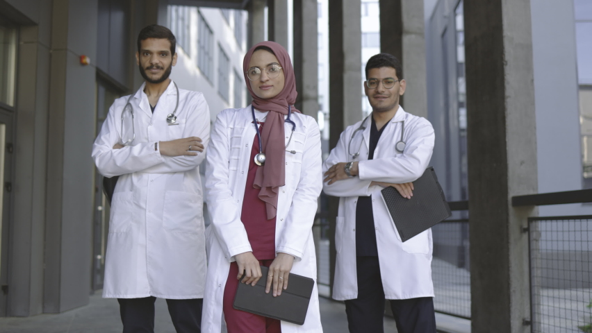 Front view of high-skilled team of three young confident Saudi Arab doctors, standing outside the hospital and looking at camera. Pretty Muslim lady in hijab stands in front of her two male colleagues | Shutterstock HD Video #1077225704