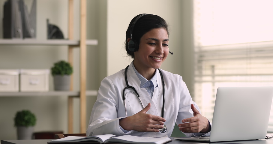 Indian female general practitioner give medical support to patient distantly use video call app, look at computer screen wear headset talk to client makes speech. Remote medicine consultation concept Royalty-Free Stock Footage #1077230915
