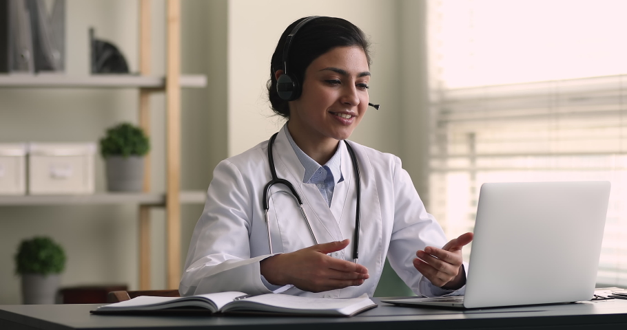 Indian female general practitioner give medical support to patient distantly use video call app, look at computer screen wear headset talk to client makes speech. Remote medicine consultation concept | Shutterstock HD Video #1077230915