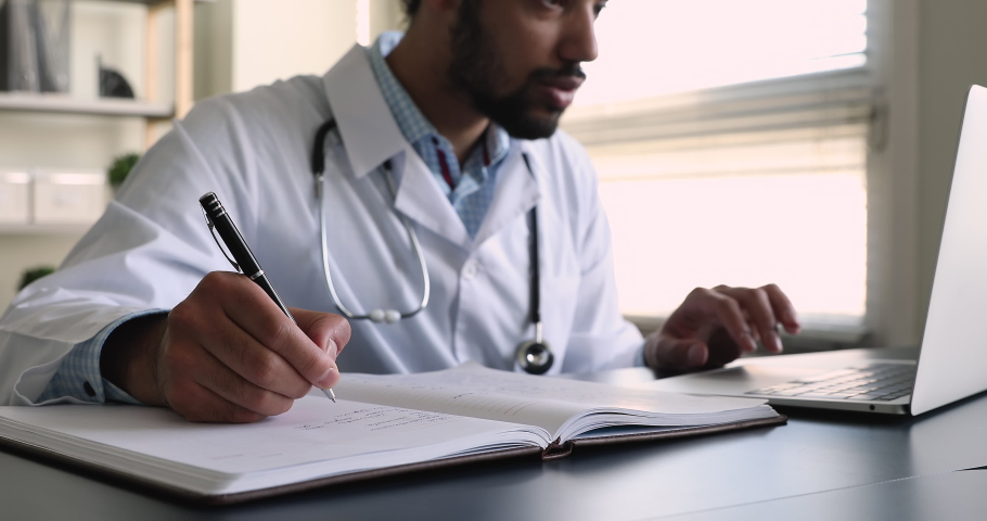 Close up African male medical worker general practitioner in uniform sit at desk holds pen take notes in medical casebook journal, use laptop check illness history, make prescription, workflow concept Royalty-Free Stock Footage #1077230963