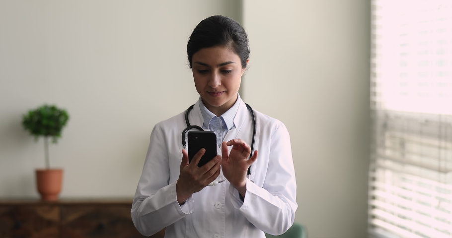 Medicine and modern tech, medical virtual app usage concept. Indian ethnicity woman doctor in white uniform standing in clinic office holding smartphone typing answer to patient, take break have fun Royalty-Free Stock Footage #1077230966