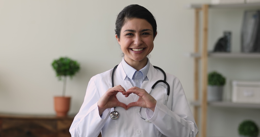 Portrait of happy Indian woman medic cardiologist in white uniform smile look at camera show love sign gesture with fingers. Cardiology, heart check up, healthcare, medical insurance, medicare concept | Shutterstock HD Video #1077230975