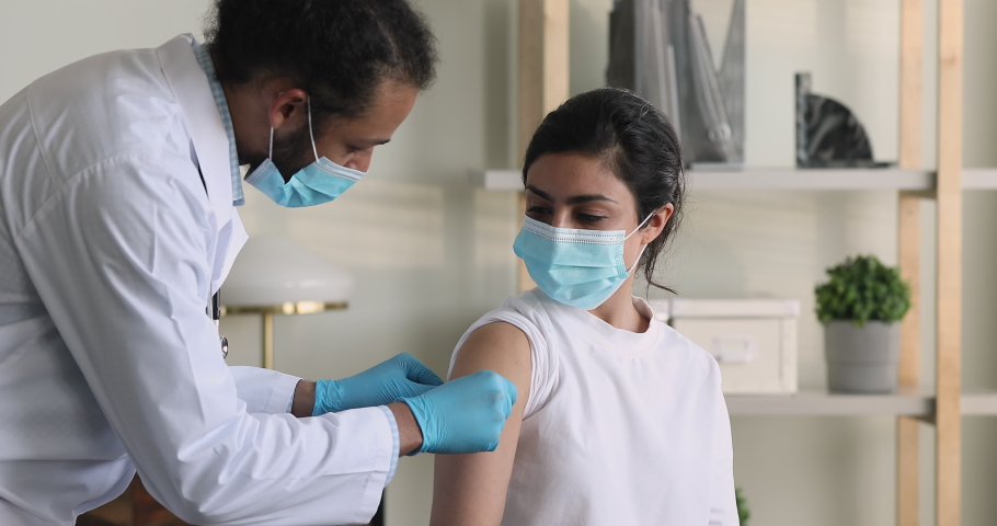 African male doctor wear surgical face mask protective gloves due precaution measures puts adhesive bandage on Indian female patient shoulder after injection against corona virus. Vaccination concept Royalty-Free Stock Footage #1077230996