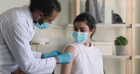 African male doctor wear surgical face mask protective gloves due precaution measures puts adhesive bandage on Indian female patient shoulder after injection against corona virus. Vaccination concept