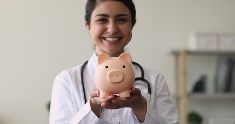 Close up young Indian female therapist wearing white coat holding piggy bank box pose in clinic office. Healthcare services expenses, health medical insurance cover, money savings and costs concept