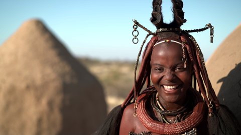 Happy young Himba woman smiling, standing next to hut in traditional Himba village near Kamanjab in northern Namibia, Africa. 