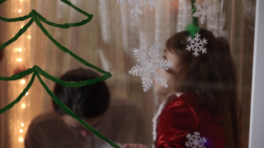 Authentic cute mom and two small preschool minor children siblings girl and boy 2-4 years old draw Christmas tree on window. young woman with children glues snowflakes. Xmas, New Year, winter concept.