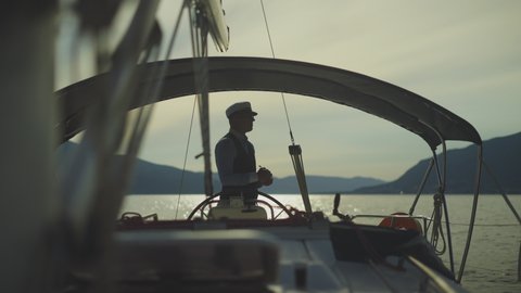 Man standing at steering wheel and lighting pipe. Silhouette of sailor in yacht cockpit. Summer vacation at sea, sailing and travelling concept.