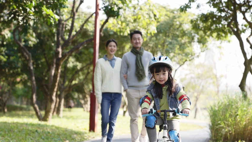 Asian little girl riding bike in park with parents walking watching in background