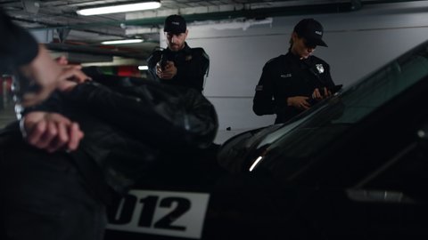 Police team arresting criminal at underground parking. Male cop handcuffing suspect at patrol car. Police officer aiming robber with gun. Policewoman using digital tablet for work