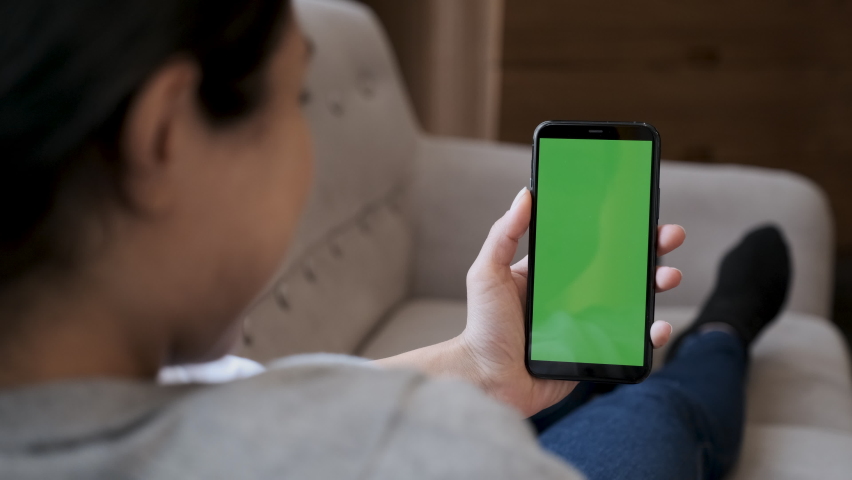 Over shoulder shot of caucasian woman holding a green screen mobile phone or smartphone in both hands, in a modern room. Close up view Royalty-Free Stock Footage #1077253817