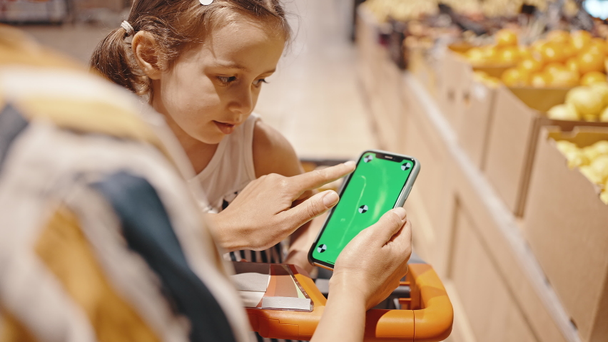 Close-up of mom and daughter in the supermarket while shopping, looking at the smartphone screen, leafing through the pages and choosing groceries. Chromakey on the phone screen. Royalty-Free Stock Footage #1077254693