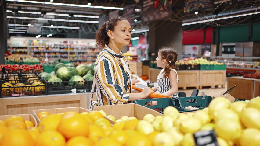 Small daughter sitting in a supermarket trolley. Lovely family, mother and little daughter buy green and red apples in the hypermarket in the fruit section. Vegetarianism and healthy eating concept. | Shutterstock HD Video #1077254714