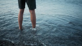 Caucasian man in shorts is standing ankle deep in water in shallow water of pebble beach. 4K slow motion video of feet in water outside close up. Small sea wave rolls over bare legs.