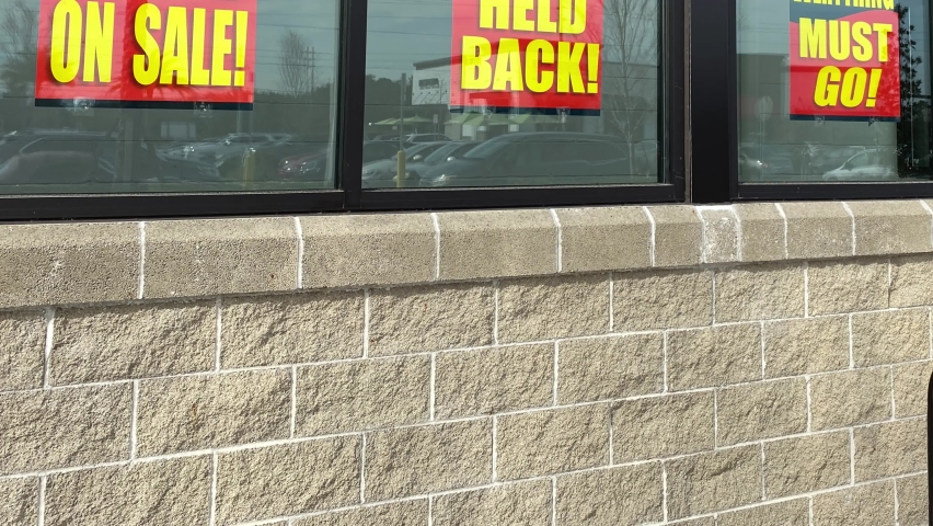 Panning up on signs in the window of a retail store going out of business that say the entire store on sale, everything must go and nothing held back.