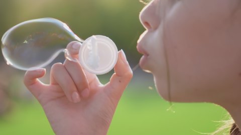 Cropped bokeh shot of preteen girl blow soap bubbles outdoors. Close up of caucasian child having fun and blowing soap bubbles in summer park