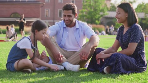 Multiethnic parents and adopted daughter sitting on lawn in park and talking. Portrait of happy diverse family with preteen girl relax together on green grass outdoors