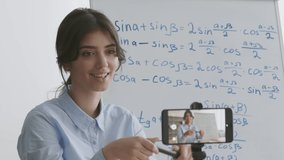 Useful blog for teens. Young smart woman recording video on smartphone, explaining mathematics formulas written on whiteboard to vlog subscribers, slow motion