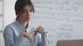 E-learning concept. Close up portrait of young professional mathematics teacher video chatting with student, explaining math formulas written on whiteboard behind her, slow motion