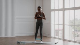 Beautiful Fit Black Woman In Sportswear Exercising With Resistance Loop Band At Home, Sporty African American Female In Wireless Headphones Lifting Leg While Standing On Yoga Mat, Slow Motion Footage