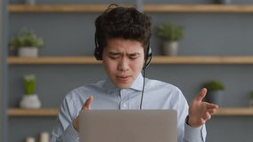 Virtual business meeting. Close up portrait of young cheerful asian guy wearing headset video chatting with coworkers via laptop, discussing project details, sitting at office, slow motion