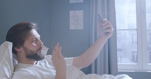 Bearded Caucasian man is lying in bed and says hello to her interlocutor, while making a video call. Beautiful young male waves his hand goodbye, while talking on the phone.