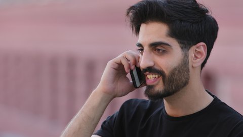 Happy smiling bearded indian guy holding cellphone at ear talking with friend. Millennial male student communicates with girlfriend. Man discuss business, calls taxi or make order of goods on phone