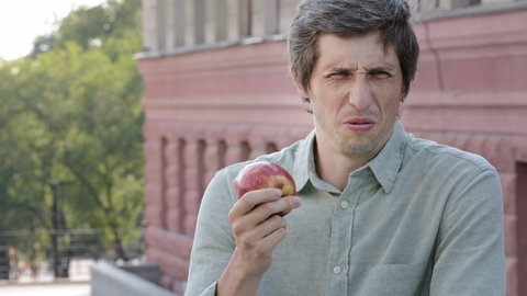Young adult male shopper customer wants to eat delicious juicy ripe apple, sniffs, makes grimace of disgust. Fruit has bad smell, is spoiled, rotten, nitrates used, unsuitable for eating. Unfair trade