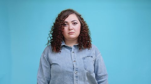 Body shaming. Obesity problem. Weight discrimination. Overweight struggle. Sad plus size obese woman with arms crossed offended by haters harassment isolated on blue background.