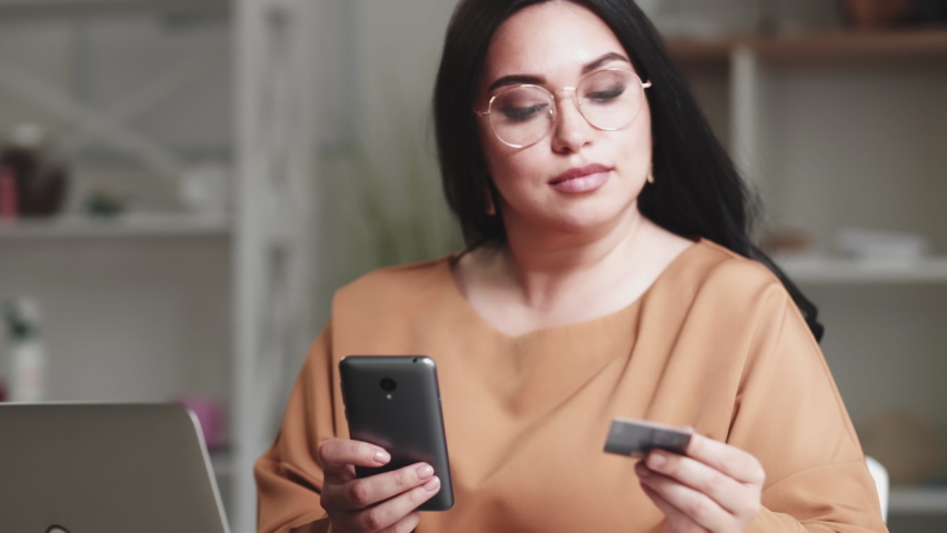 Internet payment. Online shopping. Banking service. Body positive. Concentrated cheerful obese overweight woman using phone credit card enjoying transferring in ebank app at home. Royalty-Free Stock Footage #1077268913