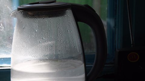 Sunlight shines in water inside teapot with blurry condensation drops on glass surface. Turbid water waving inside old transparent kettle in kitchen and making sun glares