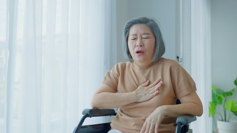Asian Senior old disabled woman suffer from heart attack on wheelchair. Elder female with chest pain has difficulty breath clutching her chest from acute pain. Hospital Healthcare and medical concept.
