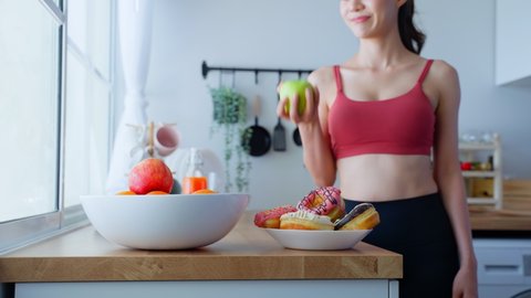 Asian attractive active woman pick up a green apple in kitchen at home. Beautiful sport girl in sportswear desire to chose fruit and ignore sweet donut enjoy eat healthy food for health after exercise