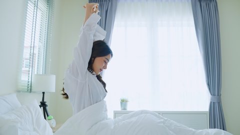 Asian beautiful girl in pajamas wake up in the morning with happiness. Attractive young woman smiling, feel happy and relax then stretching body after getting up from sleep on bed in bedroom at home..