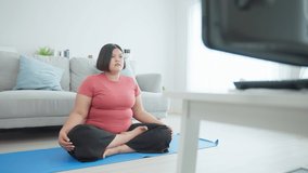 Asian young oversize woman doing yoga exercise in living room at home. Attractive plus size girl workout by follow Pilates instruction video from online trainer on television to lose weight in house.