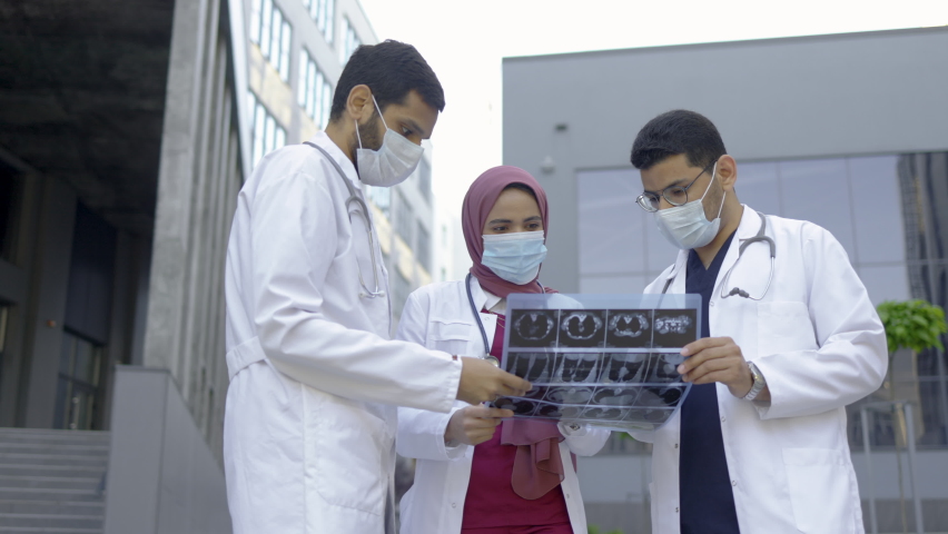 Team of three young diverse experts doctors looking at MRI picture in front of modern clinic outdoors. Pretty Muslim lady in hijab explains the results of x-ray tomography to her male colleagues | Shutterstock HD Video #1077271193