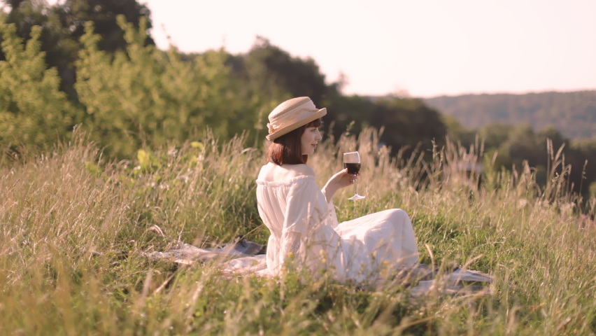 Pretty 30-aged European lady, wearing white dress and stylish straw hat, sitting on the blanket outdoors in the field, drinking red wine and enjoying wonderful nature ladscape