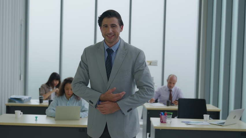 Smart Latino man standing at modern office coworking space. Portrait of young male boss manager looking to camera and gesture to celebrate joyful. Blurred employee various nationalities background.
