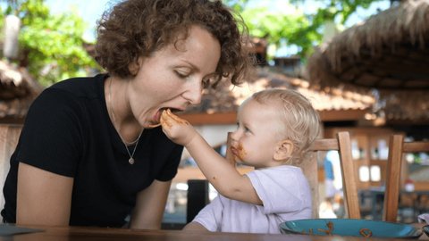 Caucasian baby feed her young mother tomato pasta with hands. Family time on lunch on a vacation