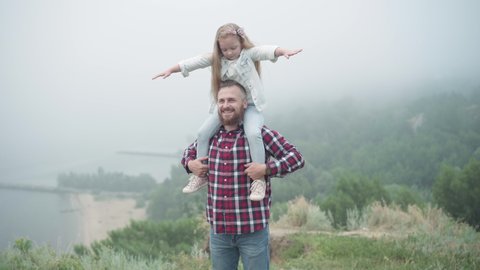 Happy smiling young bearded man stretching hands holding girl on shoulders talking looking away. Portrait of excited Caucasian father enjoying trip with cute little daughter