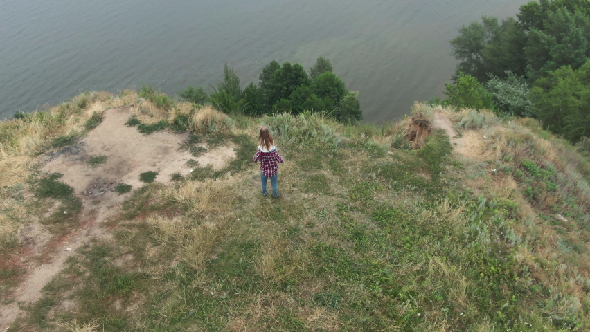Little Caucasian girl with long hair sitting on shoulders of man standing on cliff over river in mountains. Wide shot of daughter and father admiring nature outdoors. Live camera zoom out